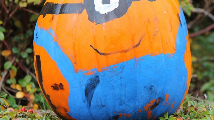 Pumpkin painted like a minion from the Despicable Me movies. The pumpkin is brigh orange with blue overalls and a single eye with goggles painted on.