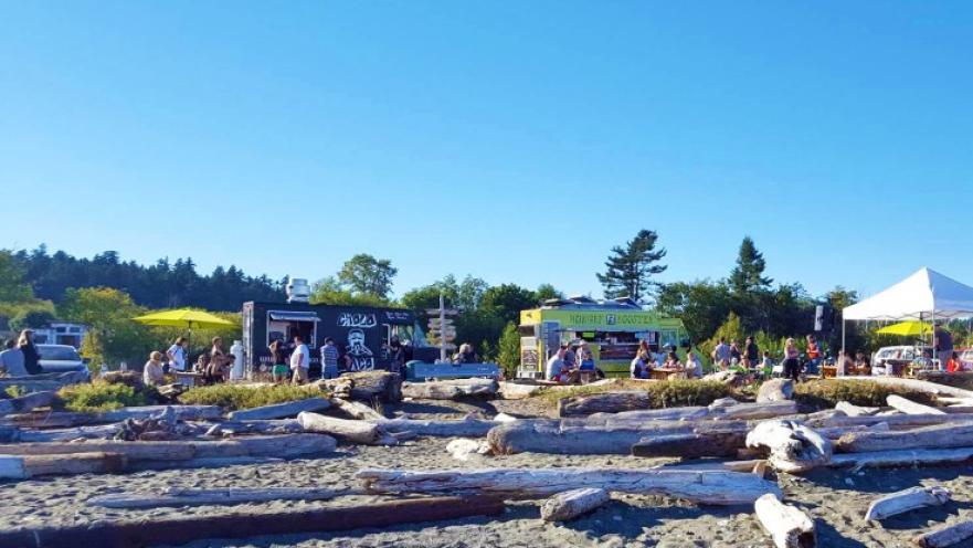 Large driftwood logs on sandy beach. There are some food trucks in the background.