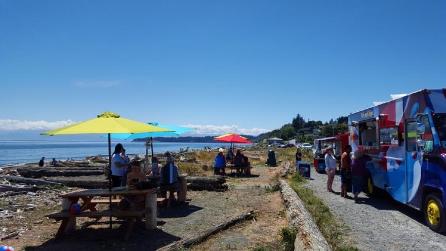 Row of picnic tables near the beach with colourful umbrellas. There are some food trucks near the picnic tables.