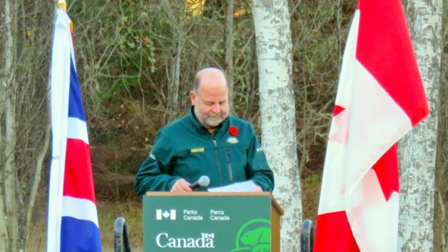 Man standing in podium flanked by the United Kingdom and Canadian flags.