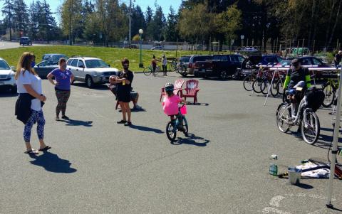 Child riding a bike at a community event in Colwood with adults standing nearby, some wearing masks.