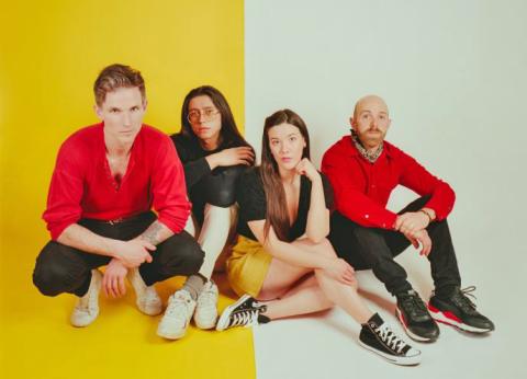 Four band members sitting against a backdrop split into yellow and white halves. Two men, one in a red shirt and one bald in a red jacket, sit on the yellow side. Two women, one in glasses wearing a black shirt and the other in a black top and yellow skirt, sit on the white side.
