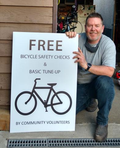 A man kneeling and holding a sign that reads "Free Bicycle Safety Checks & Basic Tune-Up by Community Volunteers," next to a symbol of a bicycle.