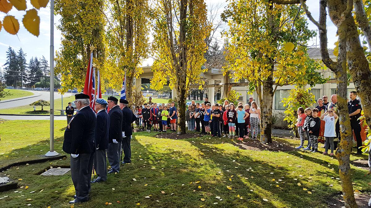 A Remembrance Day ceremony with veterans standing in uniform holding flags, while a group of children and other spectators watch.
