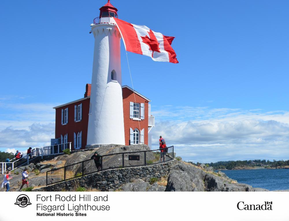 The red and white Fisgard Lighthouse with a large Canadian flag flying, located on a rocky outcrop overlooking the water at Fort Rodd Hill, a National Historic Site in Canada, under a clear blue sky.