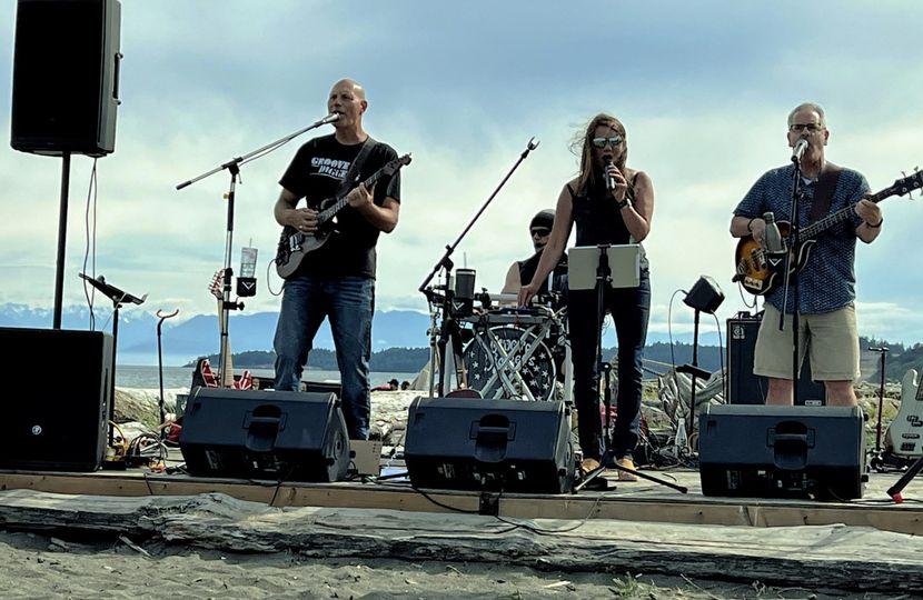 Band performing on an outdoor stage at the beach, with the ocean and mountains in the background.