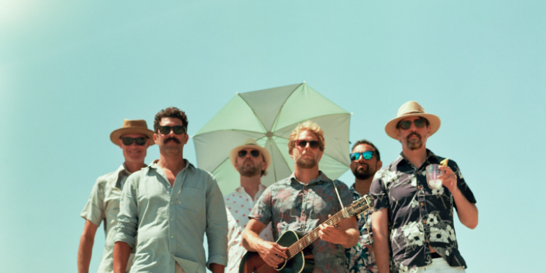 Six men in casual summer clothing standing together, one holding a guitar and another with a drink, against a clear blue sky.