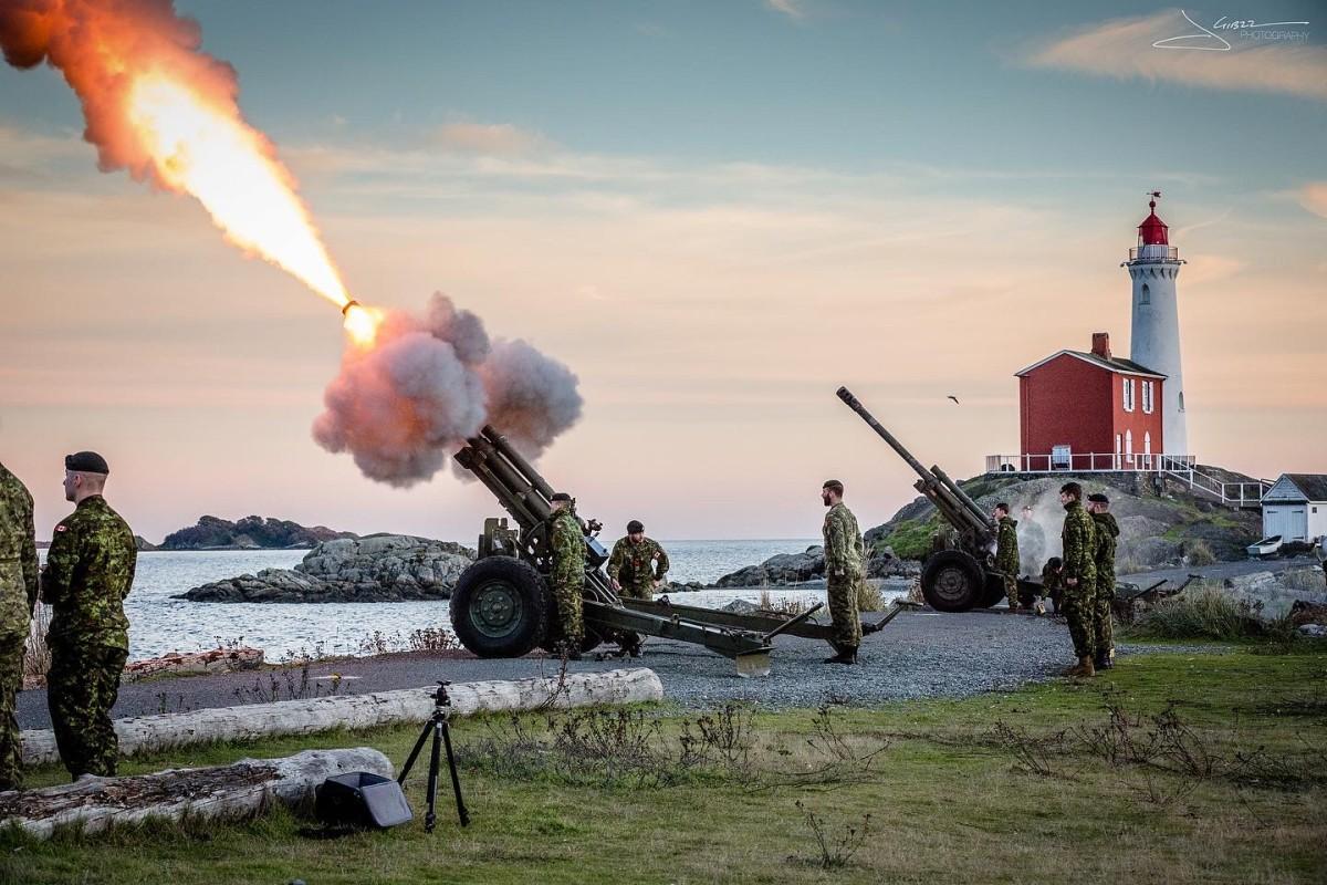 Canadian military personnel firing artillery cannons with smoke and flames near a lighthouse by the ocean during sunset.