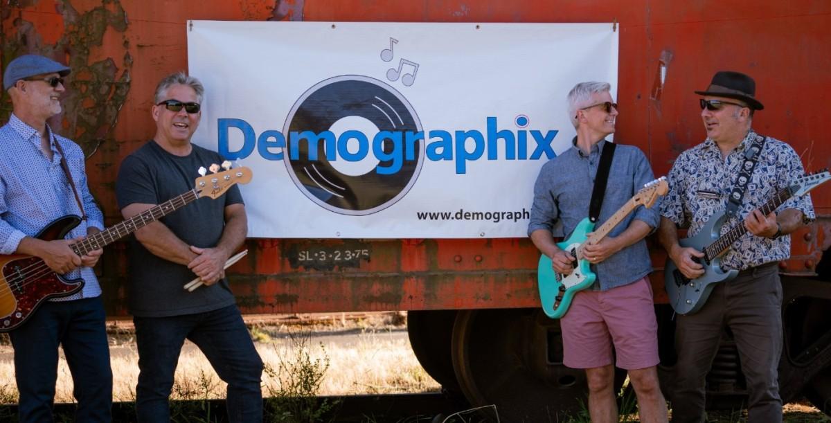 Four men from the band Demographix standing in front of a banner with their name. They are holding guitars and smiling, with an outdoor, rustic background.