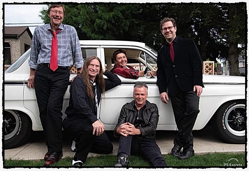 Five men posing with a vintage white car. Three are standing, one is sitting on the car door, and one is sitting on the ground in front. They are dressed in casual and semi-formal attire.