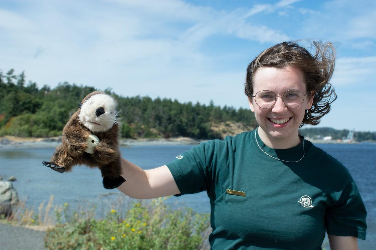 Woman holding a sea otter puppet and smiling by the waterfront with a forested background.