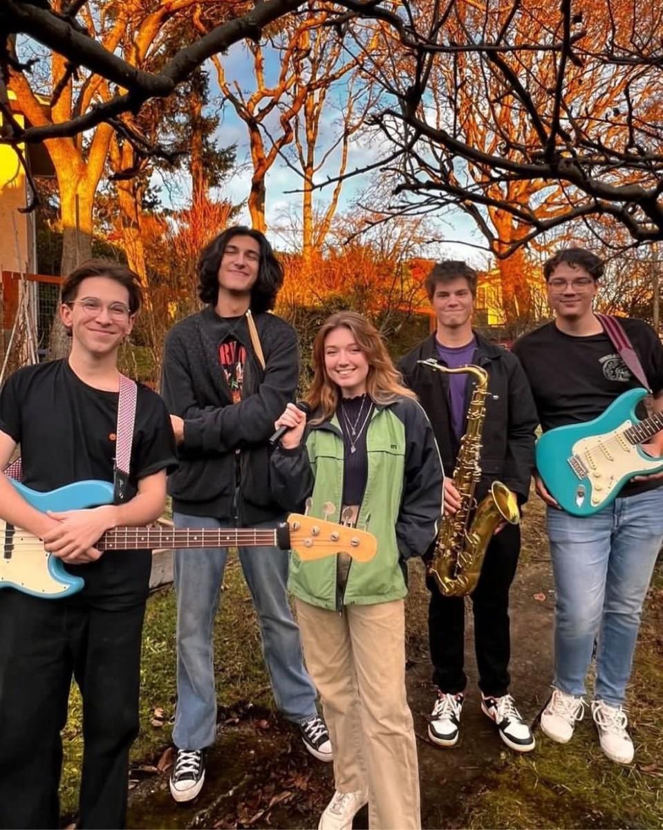 A group of five teenagers smiling and standing outdoors with musical instruments, including guitars and a saxophone, with trees in the background during sunset.